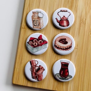 Könnte beinhalten: Satz mit 10 runden, weißen, keramischen Ansteckknöpfen mit verschiedenen Illustrationen. Die Illustrationen zeigen einen Teebeutel, eine Teekanne, einen Keks, eine Tasse Tee und eine Schachtel Kekse.