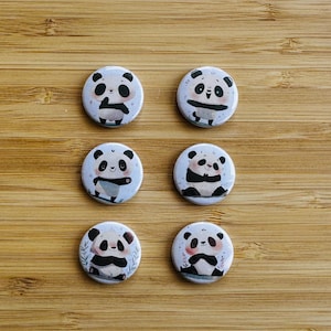 Könnte beinhalten: Sechs runde Anstecknadeln mit verschiedenen Illustrationen von Pandas. Jeder Panda ist in einer anderen Pose dargestellt, einige halten Bambus und andere lächeln. Die Nadeln sind weiß mit blauem Hintergrund und haben einen schwarzen Rand.