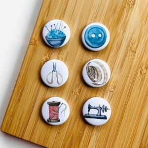 Peut inclure: Ensemble de six boutons ronds sur le thème de la couture. Chaque bouton présente une illustration à l'aquarelle différente : un coussin à épingles, un bouton bleu, des ciseaux, un mètre ruban, une bobine de fil et une machine à coudre.