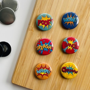 Op de afbeelding: Zeven kleurrijke ronde knoppen met een stripboekthema. De knoppen bevatten woorden als "POOF", "WOW", "BANG", "SPLASH", "WHAM" en "CRASHHH!" in vetgedrukte witte letters op een rode, blauwe of gele achtergrond.
