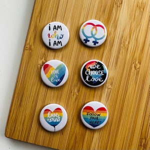 Op de afbeelding: Zes ronde regenboogkleurige knoppen met verschillende berichten. De berichten zijn "Ik ben wie ik ben", "Liefde is liefde", "We kiezen voor liefde", "Liefde wint", "Volg je hart" en een symbool van twee in elkaar grijpende cirkels.