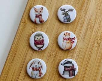 May include: Six round pins with different woodland animals wearing winter scarves. The animals are a fox, a moose, a hedgehog, a squirrel, a rabbit, and a badger.