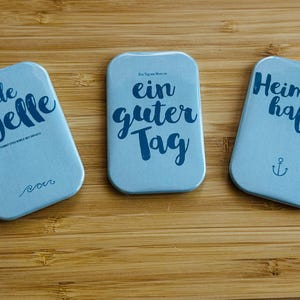 Könnte beinhalten: Drei blaue rechteckige Magnete mit weißem Text. Der erste Magnet sagt "jede Welle" mit einem Wellensymbol darunter. Der zweite Magnet sagt "Ein Tag am Meer ist ein guter Tag". Der dritte Magnet sagt "Heimathafen" mit einem Ankersymbol darunter.