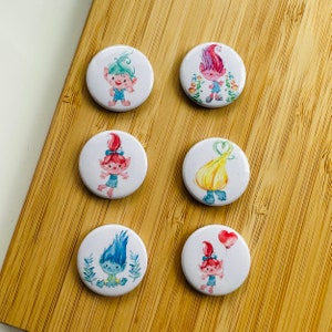 Könnte beinhalten: Sechs runde, weiße, knopfartige Anstecker mit bunten Comic-Bildern von Trollen. Die Trolle haben blaues, grünes, rosa und gelbes Haar. Einige Trolle tragen Kleidung, andere nicht. Die Anstecker sind perfekt, um Ihren Gegenständen einen Hauch von Fantasie zu verleihen.