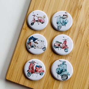 Könnte beinhalten: Sechs runde weiße Anstecknadeln mit bunten Illustrationen von Vintage-Rollern. Jede Nadel zeigt ein anderes Rollerdesign mit verschiedenen Farben und Mustern.