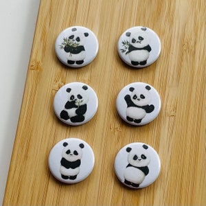 Peut inclure: Six boutons ronds blancs avec des illustrations de pandas noirs et blancs. Les pandas sont dans différentes poses, y compris tenant du bambou, assis et debout.