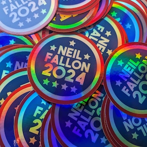 May include: A collection of circular, holographic stickers. Each sticker features the text "NEIL FALLON 2024" in white on a blue background, with stars. The stickers have a red border and a rainbow sheen.