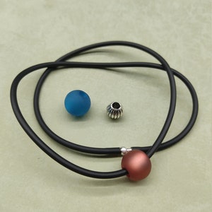 Könnte beinhalten: Eine schwarze Schnurkette mit einer kupferfarbenen Perle und einer silbernen Perle. Die Kette liegt auf einer hellgrünen Oberfläche.