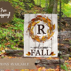 May include: A white wooden garden flag with a fall wreath featuring a brown letter "R" in the center. The wreath is made of brown branches and yellow leaves. The text "welcome fall" is written in brown below the wreath.