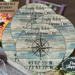 May include: A round wooden sign with a blue and white distressed woodgrain background. The sign features a compass rose with the text "Empty Pockets Saratoga Springs, NY" and the coordinates "43°02′55″ N 73°43' 27" W".