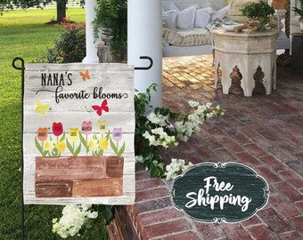 May include: A white wooden garden flag with the text "Nana's favorite blooms" and a floral design featuring colorful flowers and the names of family members. The flag is hanging on a metal pole.
