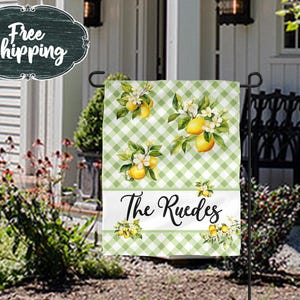 May include: A rectangular garden flag featuring a light green and white gingham pattern with illustrations of yellow lemons and white blossoms. A horizontal white band across the lower section displays the black cursive text The Ruedes.