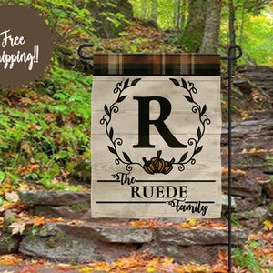 May include: A rectangular garden flag featuring a black plaid top border and a light wood grain background. The design includes a large black letter R centered within a leafy wreath, a small pumpkin illustration, and the text The RUEDE Family.