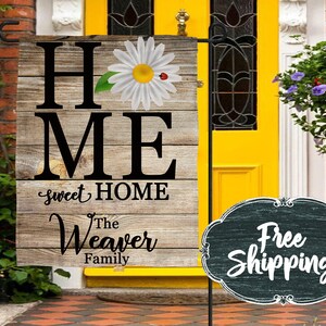 Op de afbeelding: Een houten bord met de woorden "Home Sweet Home" en "The Weaver Family" met een madeliefje en lieveheersbeestje afbeelding. Het bord wordt getoond voor een gele deur met een bloempot.