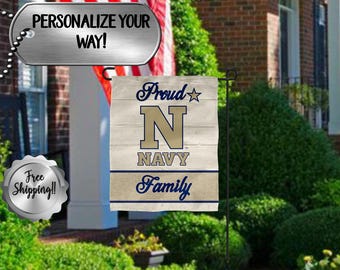 Puede incluir: Una bandera de jardín con un diseño azul marino y beige. La bandera presenta el texto "Proud Navy Family" con una gran "N" en el centro. La bandera se muestra en un soporte de metal negro en un jardín.