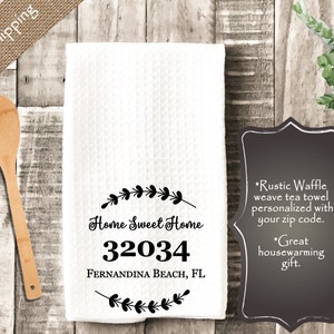 May include: White waffle weave tea towel with black text that reads "Home Sweet Home 32034 FERNANDINA BEACH, FL" surrounded by a wreath of leaves.