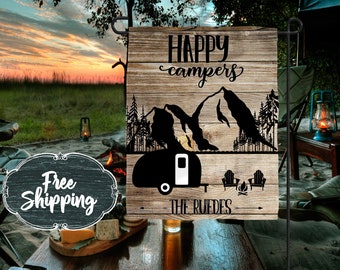 May include: A wooden camping themed flag with the text "Happy campers" and a silhouette of mountains and a camper. The flag also has the text "The Ruedes" at the bottom.
