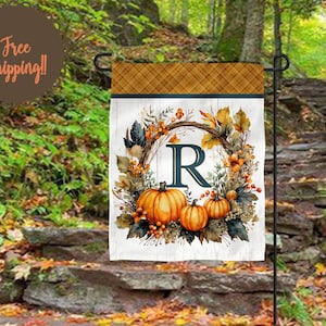 May include: A rectangular garden flag featuring a top border with a brown and orange plaid pattern. The center displays a white wood-grain background with a wreath of autumn leaves, berries, and three orange pumpkins surrounding the letter R.