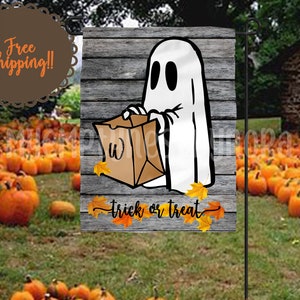 Könnte beinhalten: Ein weißer Geist, der eine braune Papiertüte mit dem Text "Trick or Treat" und Herbstblättern auf einem Holzgrund hält.