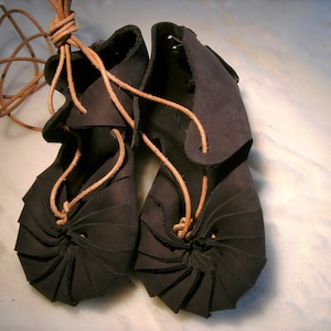 May include: A pair of dark brown leather sandals with laces. The sandals have a unique design with the leather folded and stitched to create a layered effect.