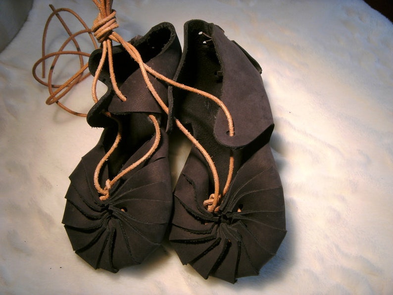 May include: A pair of dark brown leather sandals with laces. The sandals have a unique design with the leather folded and stitched to create a layered effect.