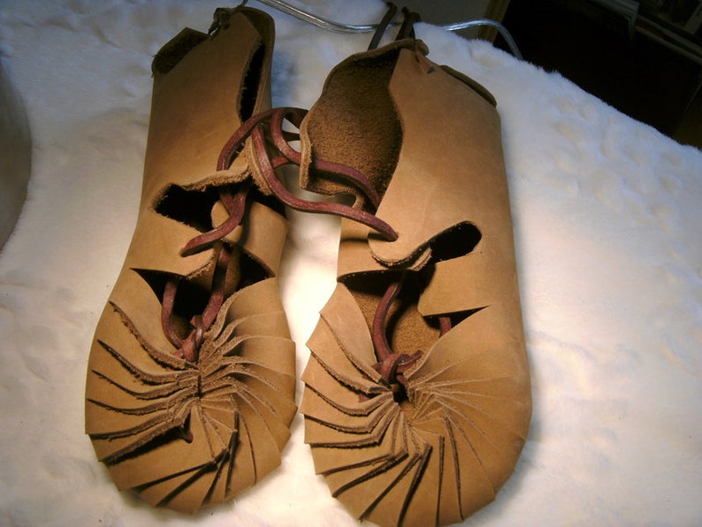 May include: A pair of brown leather sandals with a unique spiral design. The sandals are laced up the front with brown leather laces.