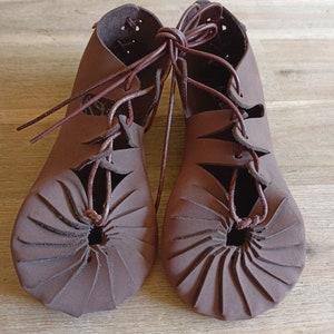 May include: A pair of brown leather sandals with laces. The sandals have a unique design with a circular pattern on the sole.