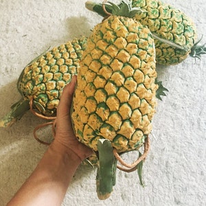 Könnte beinhalten: Drei gelbe und grüne Ananas mit einer strukturierten Oberfläche. Die Ananas sind zu einem Stapel angeordnet, wobei eine Ananas in einer Hand gehalten wird.