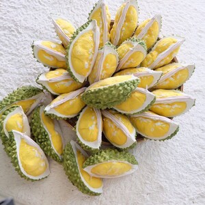 May include: A close-up of a group of durian fruit, some are whole and some are cut open, revealing the yellow flesh inside. The durian fruit are arranged in a basket and are on a white surface.