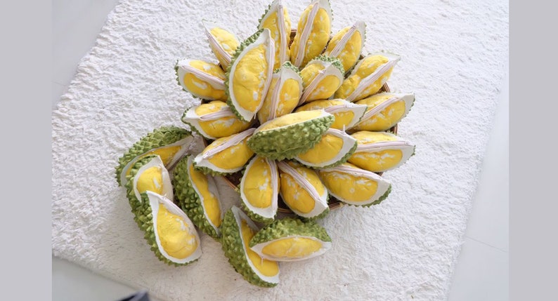 May include: A close-up of a group of durian fruit, some are whole and some are cut open, revealing the yellow flesh inside. The durian fruit are arranged in a basket and are on a white surface.