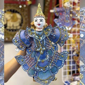May include: A blue and gold puppet with a white face and a crown. The puppet is wearing a blue and gold costume with sequins and beads. The puppet is holding a staff in its right hand. T1
