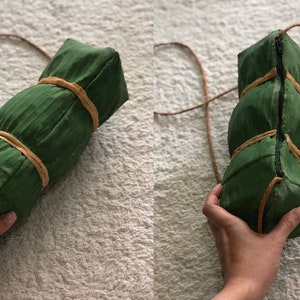 Puede incluir: Una bolsa de hoja de pl&aacute;tano verde con una correa de cuero marr&oacute;n. La bolsa est&aacute; atada con cuerda marr&oacute;n.