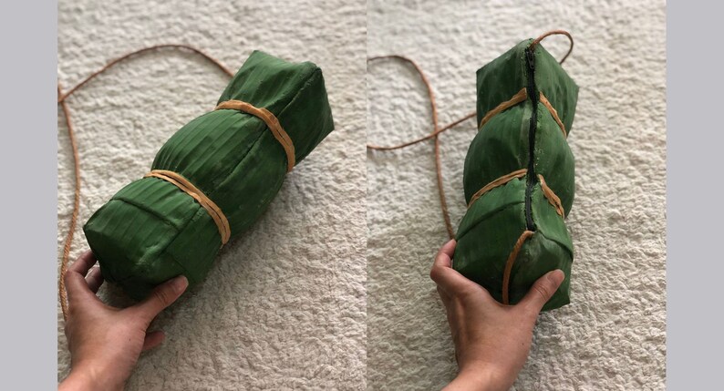 Puede incluir: Una bolsa de hoja de pl&aacute;tano verde con una correa de cuero marr&oacute;n. La bolsa est&aacute; atada con cuerda marr&oacute;n.