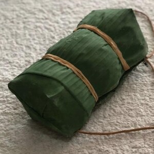 Puede incluir: Un paquete verde rectangular envuelto en hojas de pl&aacute;tano y atado con una cuerda marr&oacute;n.