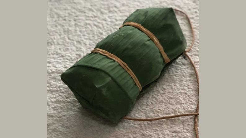 Puede incluir: Un paquete verde rectangular envuelto en hojas de pl&aacute;tano y atado con una cuerda marr&oacute;n.