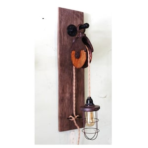 May include: A rustic wooden wall-mounted light fixture with a pulley and a caged light bulb. The pulley is made of wood and metal and has a rope attached to it. The light bulb is hanging from the rope and is encased in a wire cage.