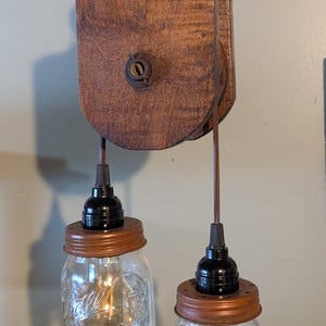 May include: Rustic, two-light pendant lamp featuring clear glass mason jars with copper-colored lids. The lamp hangs from a wooden pulley system with a metal ring. The wood has a warm, brown tone.