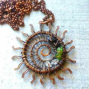 May include: Copper wire sun pendant necklace with green and black stones. The pendant is made of copper wire and features a spiral design with green and black stones.