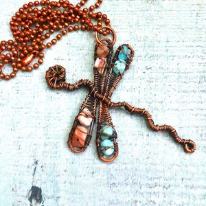 Puede incluir: Un collar con un colgante de lib&eacute;lula de alambre de cobre con acentos de piedras preciosas turquesa y rosas.