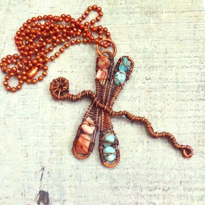 Puede incluir: Un collar de alambre de cobre con un colgante de lib&eacute;lula con piedras preciosas rosas y azules.