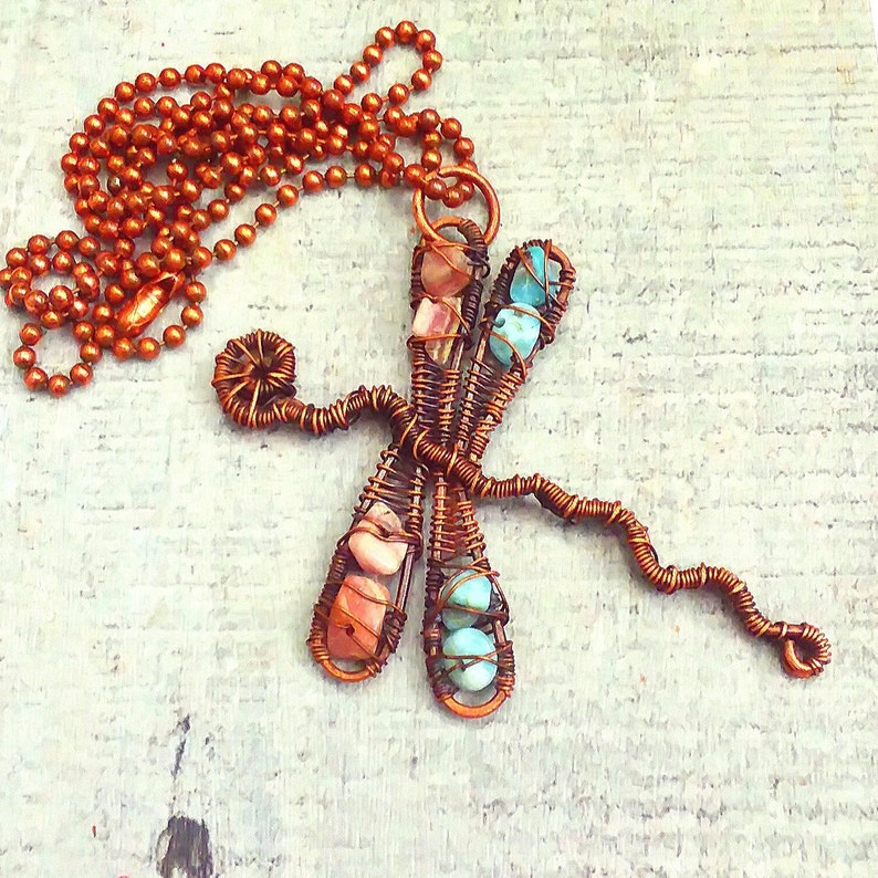 Puede incluir: Un collar de alambre de cobre con un colgante de lib&eacute;lula con piedras preciosas rosas y azules.