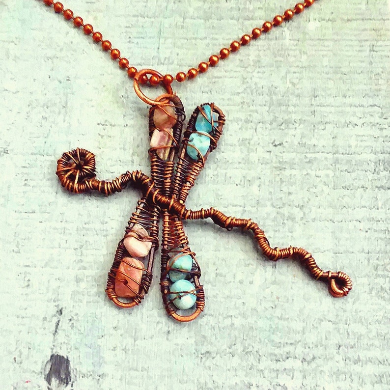 Puede incluir: Un collar de alambre de cobre con un colgante de lib&eacute;lula con piedras turquesa y rosas.