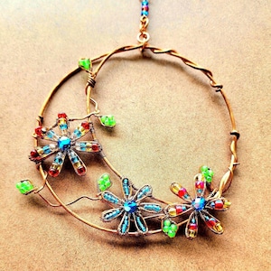 May include: A copper wire hoop with three colorful beaded flowers. The flowers are made with red, blue, green, and yellow beads. The hoop is hanging from a copper wire chain.