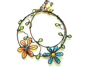 Puede incluir: Una corona de alambre con dos flores de cuentas, una naranja y una azul, con hojas verdes. La corona está hecha de alambre de cobre y está decorada con cuentas.