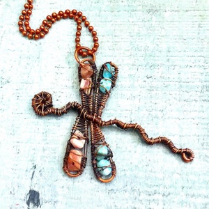 Puede incluir: Un collar con un colgante de lib&eacute;lula de alambre de cobre con piedras turquesa y rosas. El collar tiene una cadena marr&oacute;n.