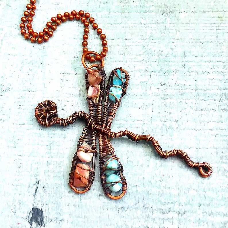 Puede incluir: Un collar con un colgante de lib&eacute;lula de alambre de cobre con piedras turquesa y rosas. El collar tiene una cadena marr&oacute;n.