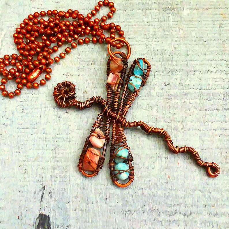 Puede incluir: Un collar con un colgante de lib&eacute;lula de alambre de cobre con acentos de piedras preciosas rosas y turquesas. El collar tiene una cadena de cobre.