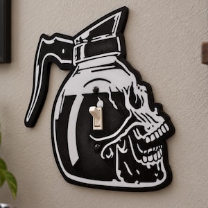 May include: A black and white light switch cover plate shaped like a coffee pot with a skull design integrated into the glass portion. The cover features a central toggle switch and is mounted on a wall, displaying a graphic illustration.