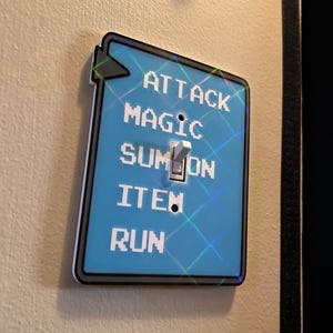 May include: A light blue light switch cover with the words "ATTACK", "MAGIC", "SUMMON", "ITEM", and "RUN" in white block letters. The switch is in the "ON" position. The cover has a black border and a holographic design.