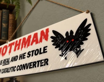 May include: A rectangular white sign hanging by a brown rope, featuring a black graphic of a winged creature with red eyes. The text in red and black reads: MOTHMAN IS REAL AND HE STOLE MY CATALYTIC CONVERTER. The sign has a smooth, glossy finish.
